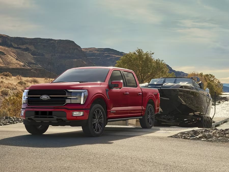 Red Ford F-150 pickup truck parked at a boat ramp, towing a sleek black boat, with mountains and trees in the background, showcasing its towing capability and rugged design.