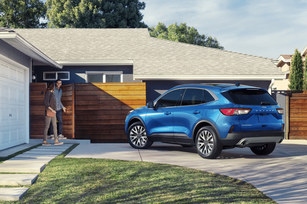 A blue SUV parked in a driveway with two people walking towards it, set in a residential neighborhood with a wooden fence and modern house design.