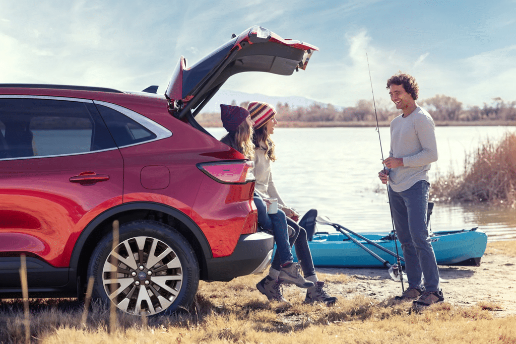 Outdoor scene featuring a red SUV parked by a lake, with a man fishing and two people sitting on the back of the vehicle. The woman is wearing a colorful beanie and the other person is holding a mug, enjoying the day. A kayak is visible in the background, highlighting a recreational adventure atmosphere.