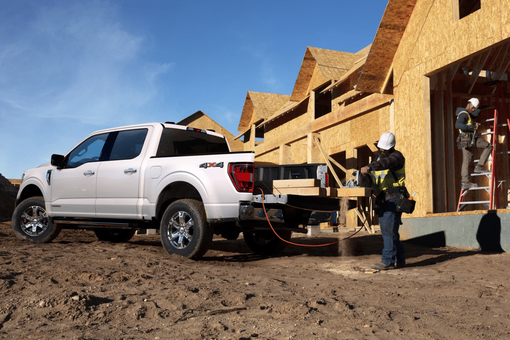 Construction workers using a white pickup truck on a building site, with one worker operating a saw on wood planks while another is seen on a ladder working on a house under construction. The background features partially built homes and a clear blue sky.
