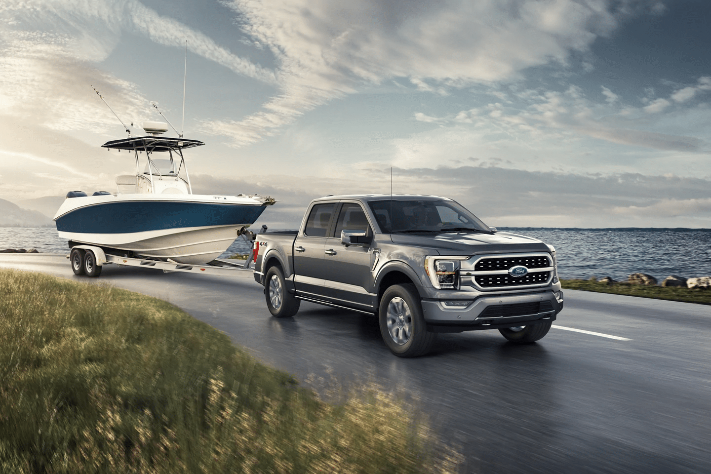 Ford F-150 towing a boat trailer along a scenic coastal road, showcasing the truck's powerful towing capabilities against a backdrop of grass and water.