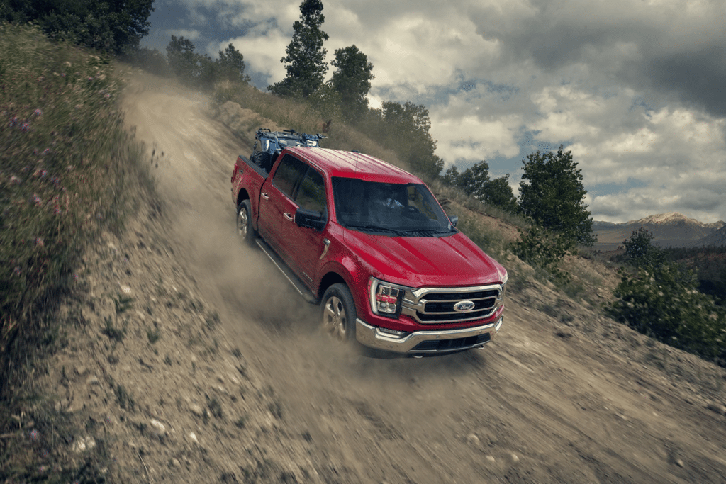Red Ford F-150 truck navigating a rugged dirt road in a scenic outdoor setting, showcasing off-road capabilities with dust clouds and rocky terrain. Ideal for adventure and utility.