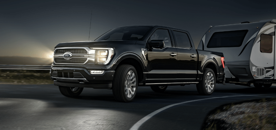 Black Ford F-150 truck towing a camper on a winding roadway at sunset, showcasing its powerful performance and towing capabilities.