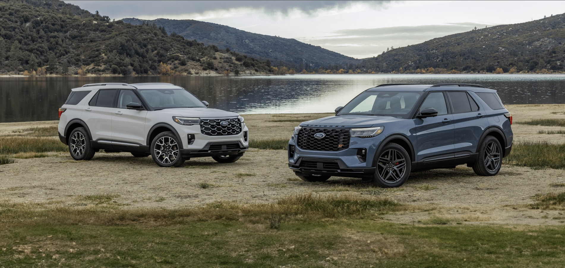 Two Ford Explorer SUVs parked on a grassy shore by a lake, with mountains in the background. One vehicle is white, while the other is blue. The scene captures a cloudy sky and serene landscape, showcasing the adventurous spirit of the Explorer lineup.