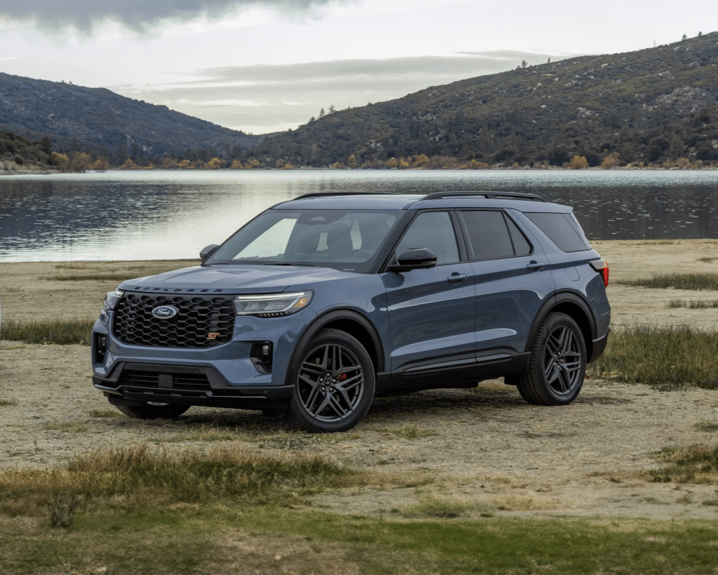 Two Ford Explorer SUVs parked near a serene lake, surrounded by mountains and greenery. The left SUV is white with dark wheels, while the right SUV is blue with sporty accents. The image highlights their sleek designs and powerful presence in a natural setting.