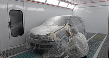 A technician in protective clothing spray paints a car inside a professional paint booth, with the vehicle covered in plastic to protect its surfaces.