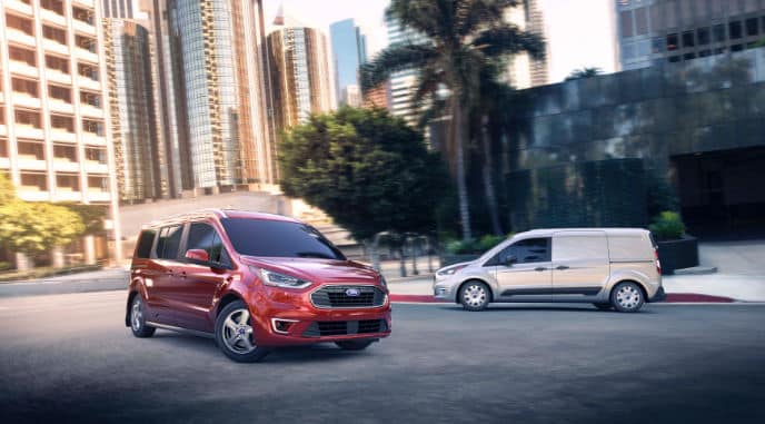 Two Ford vans, a red model and a gray model, driving on a city street lined with tall buildings and palm trees, showcasing urban versatility and style.