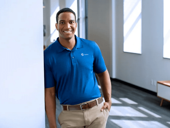 Smiling man in a blue polo shirt leaning against a wall in a bright, modern office space, showcasing a professional and approachable demeanor.