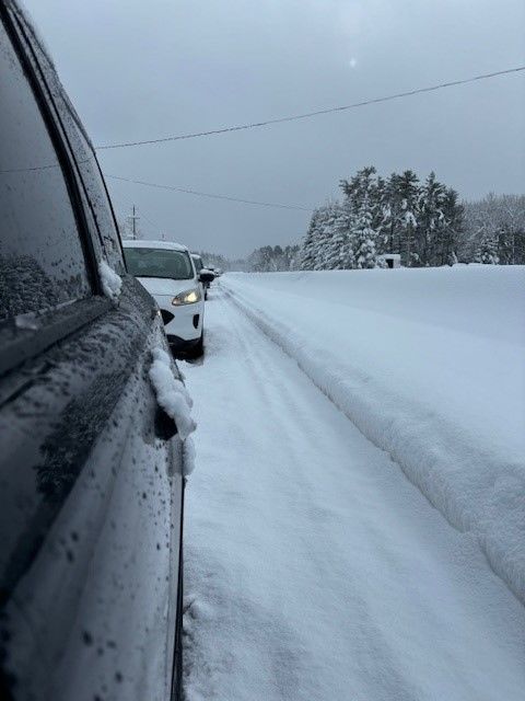 Snow-covered road with two vehicles parked on the side, surrounded by trees, under a cloudy sky. The image captures the challenges of winter weather driving conditions.