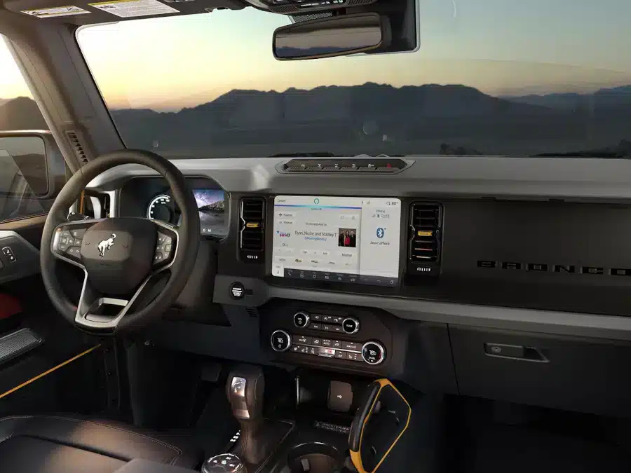 Interior view of a Ford Bronco showcasing a modern dashboard with touchscreen display, steering wheel with logo, and various controls in a scenic setting with mountains in the background at sunset.