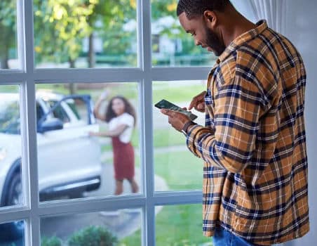 Man in a plaid shirt looking at his smartphone while standing by a window; a woman in a red dress waves from outside near a white SUV.