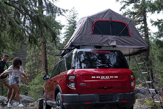 Red Ford Bronco Sport parked in a forest setting with a rooftop tent set up. Two people, a man and a woman, are climbing rocks near the vehicle, enjoying outdoor camping activities. Lush trees surround the scene, highlighting a nature escape perfect for adventure seekers.