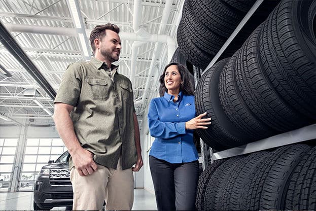 Customer and service representative discussing tire options in a modern auto service center, with a display of tires in the background and a vehicle parked nearby.